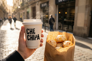 Fotografía de una bolsa de chipás artesanales de queso recién horneados y un café con leche para llevar de Che Chipá en el barrio de Gràcia.